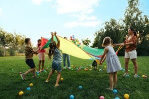Group of children and teacher playing with rainbow playground parachute on green grass. Summer camp activity